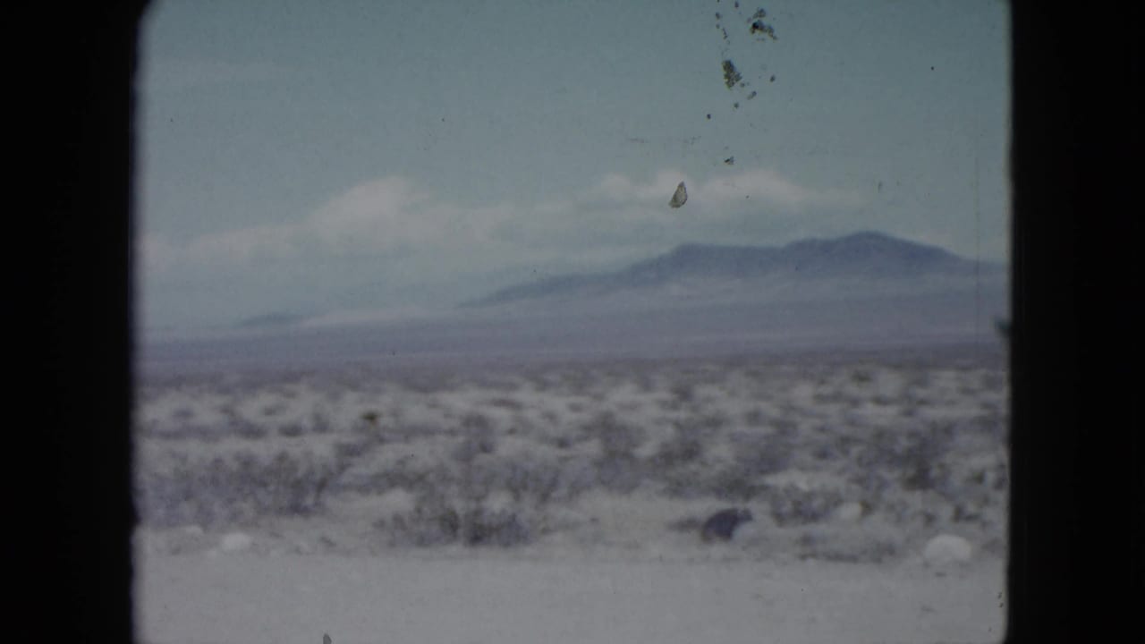 1959 Nevada home-movie still of scrubby desert terrain, pale foreground, distant mountain ridge, and soft clouds in a faded 8mm frame.