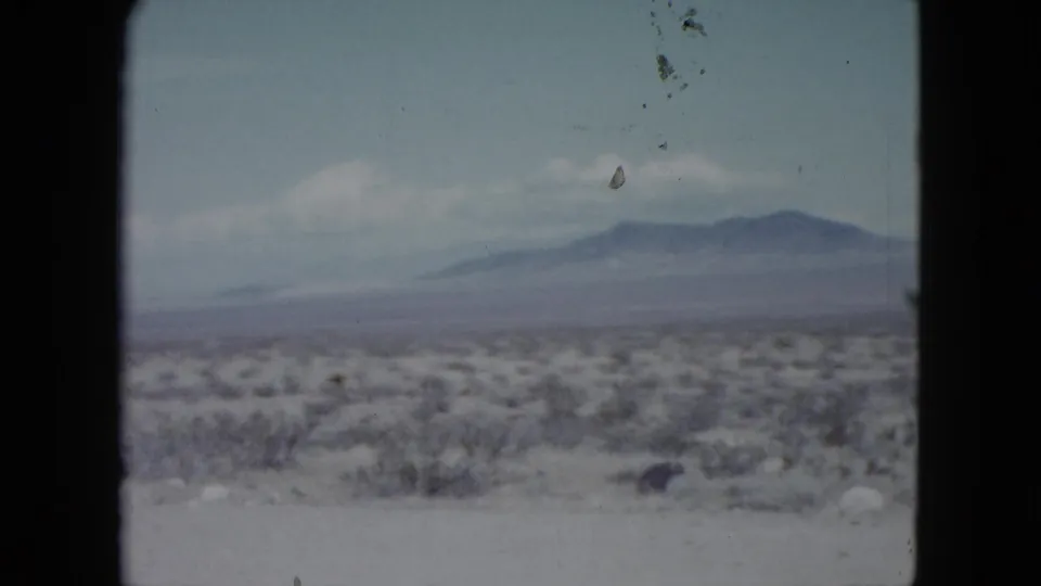 1959 Nevada home-movie still of scrubby desert terrain, pale foreground, distant mountain ridge, and soft clouds in a faded 8mm frame.