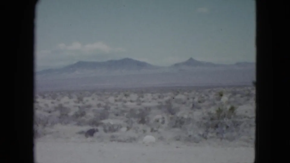 Nevada 1959 8mm frame with low desert plants, light ground, distant layered mountains, and a small pointed peak on the horizon.