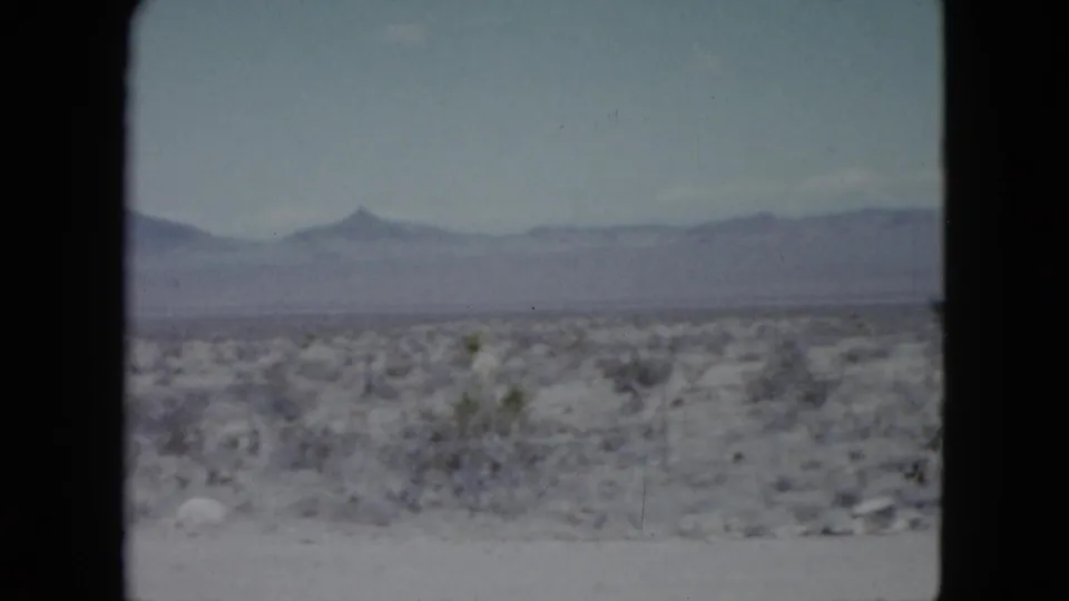 Faded 1959 Nevada 8mm still showing open desert scrub, scattered stones, a distant mountain range, and a pointed peak under blue sky.