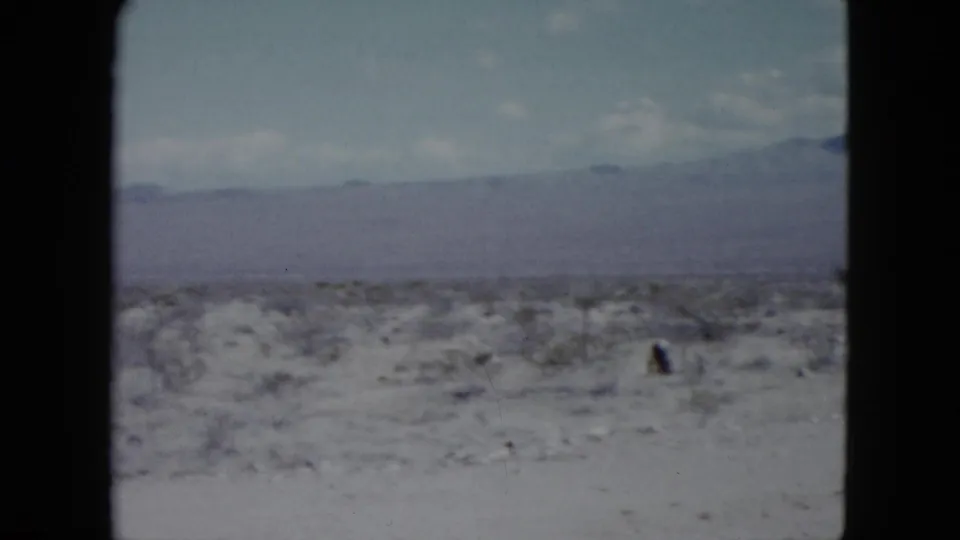 1959 Nevada desert frame with pale ground, small shrubs, soft clouds, and mountains in the background.