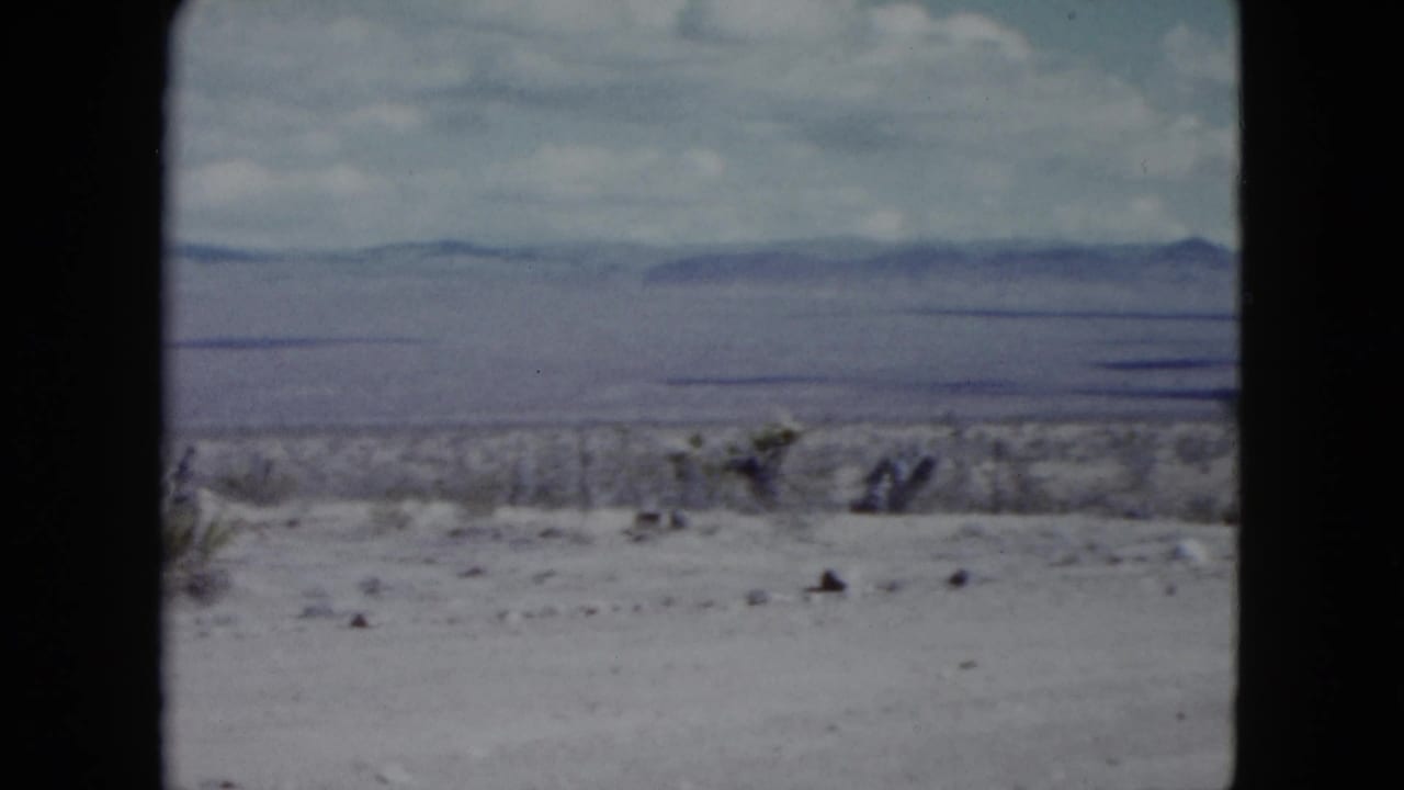 1959 Nevada desert view with low brush, a broad dry plain, and hazy mountain ridges framed by visible 8mm gate edges.