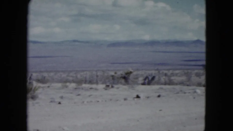 1959 Nevada desert view with low brush, a broad dry plain, and hazy mountain ridges framed by visible 8mm gate edges.