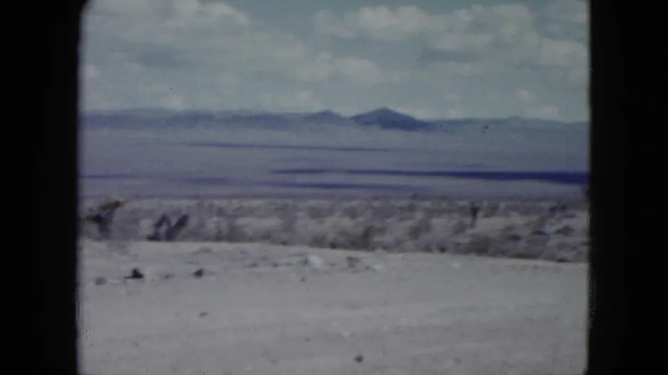 Nevada 1959 home-movie frame of open desert foreground, scattered scrub, and a low mountain skyline under a bright cloudy sky.