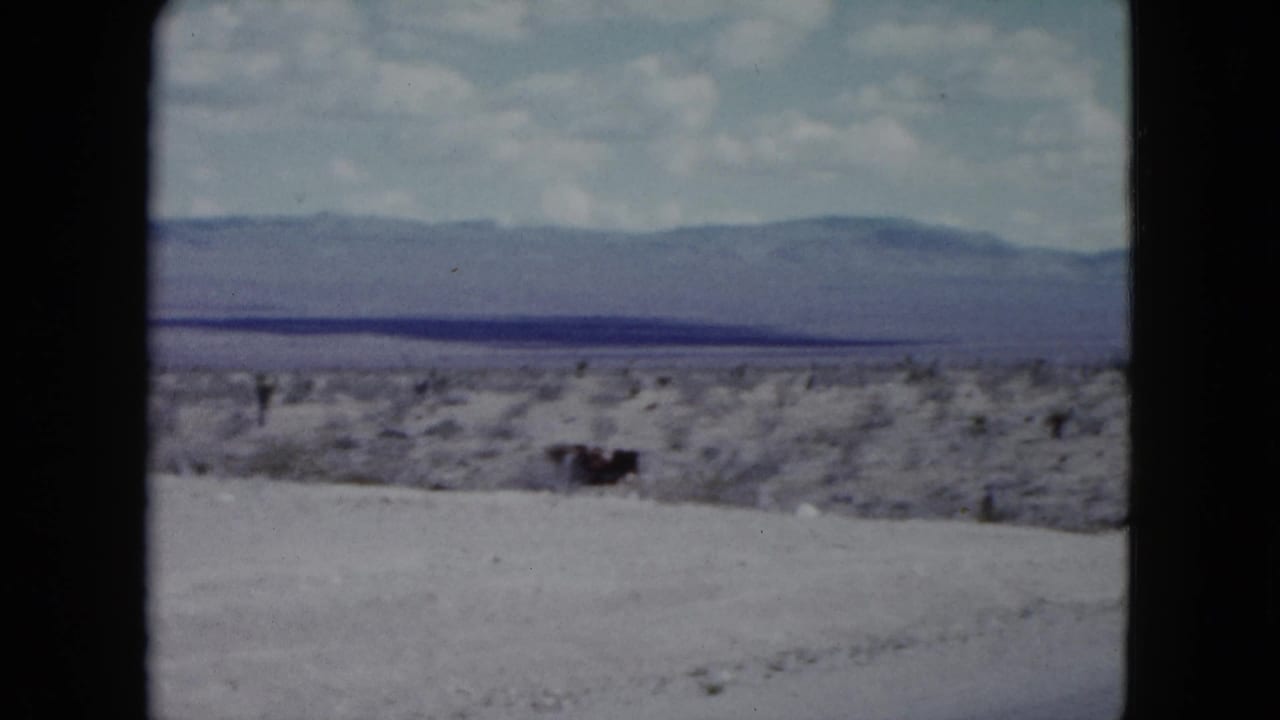 1959 Nevada home-movie frame showing scrubby desert terrain and distant mountains near a paved highway.