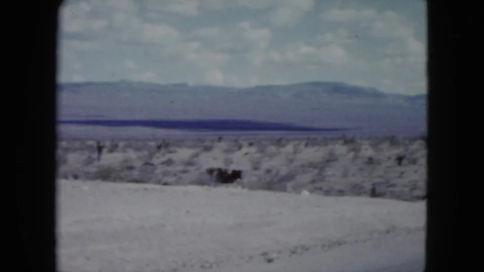 1959 Nevada home-movie frame showing scrubby desert terrain and distant mountains near a paved highway.