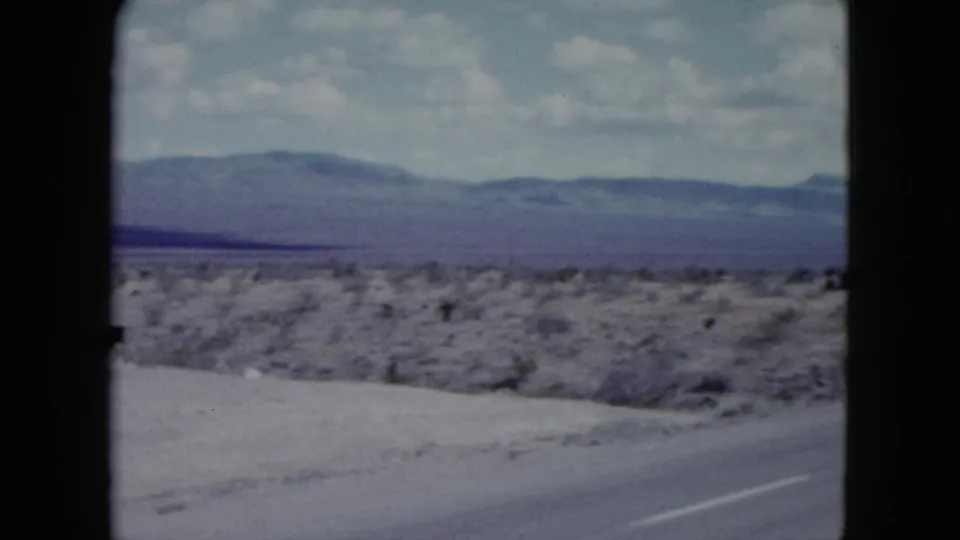 1959 Nevada 8mm frame with a paved desert highway, white road edge, scrubland, and mountains under clouds.