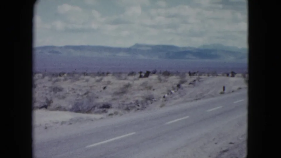 1959 Nevada roadside frame showing the highway shoulder, sparse desert brush, roadside posts, and distant mountains.