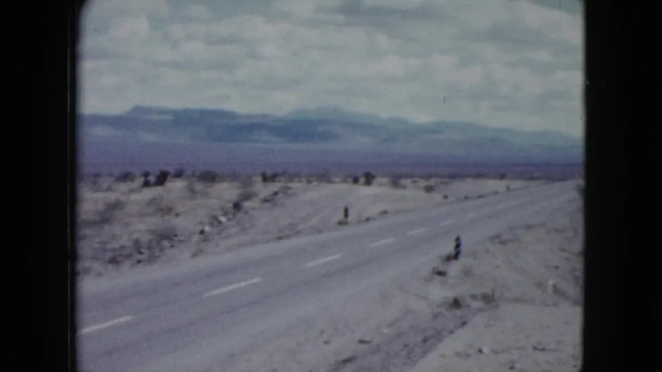 1959 Nevada home-movie frame looking along a desert highway bordered by scrub and distant mountains.
