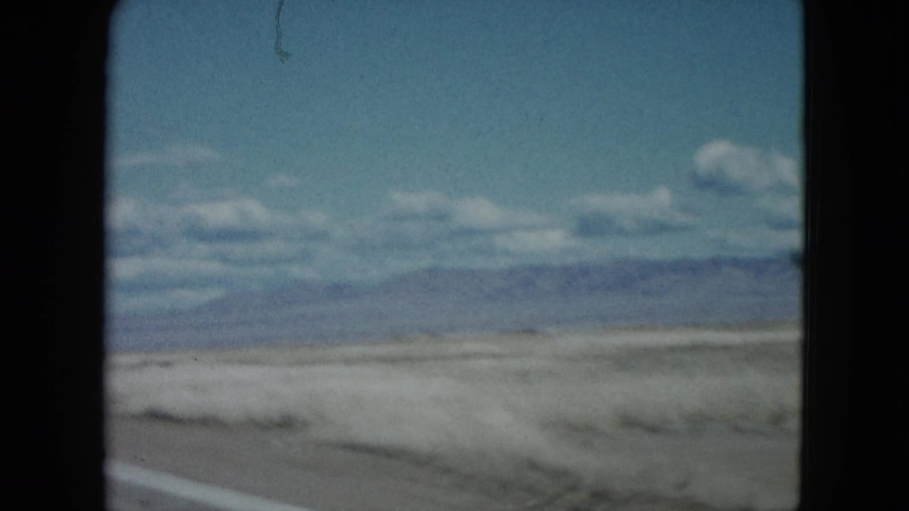 1959 Nevada roadside view from a moving camera, showing dry ground, low vegetation, distant mountains, and scattered clouds.