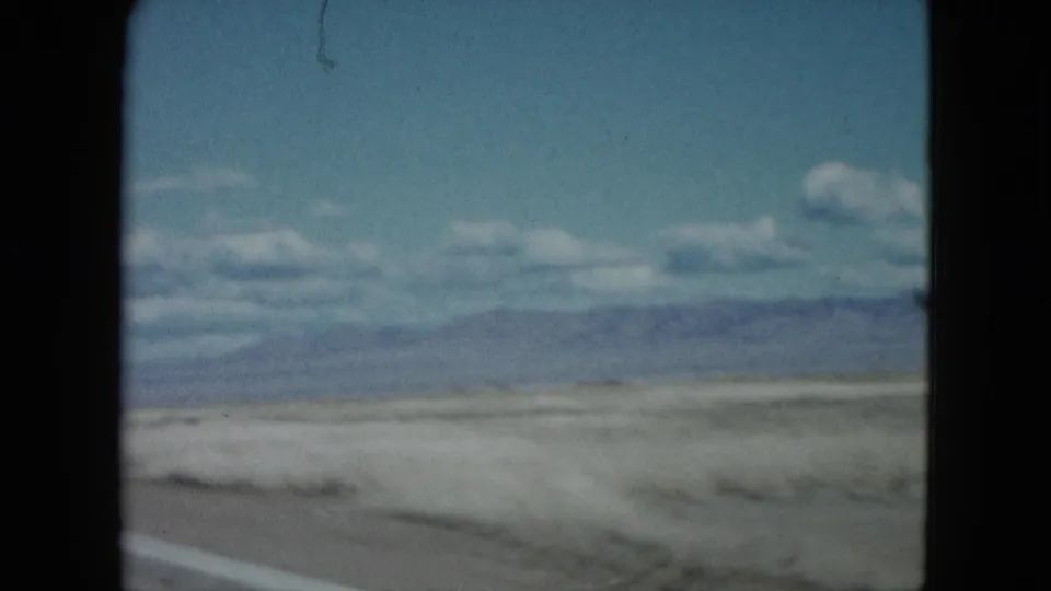 1959 Nevada roadside view from a moving camera, showing dry ground, low vegetation, distant mountains, and scattered clouds.