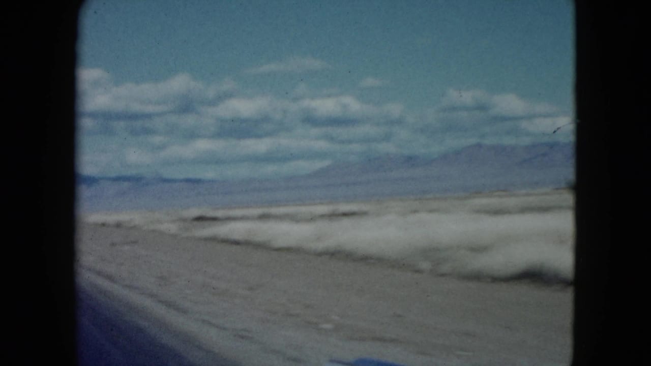 Faded 8mm frame of a Nevada desert road view, with scrubby ground, open sky, and a mountain range on the horizon.