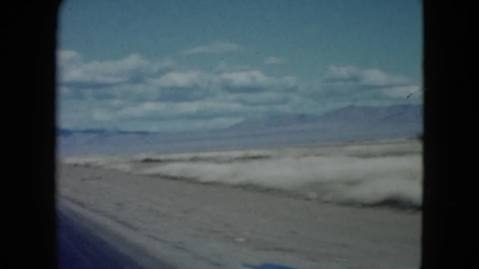Faded 8mm frame of a Nevada desert road view, with scrubby ground, open sky, and a mountain range on the horizon.