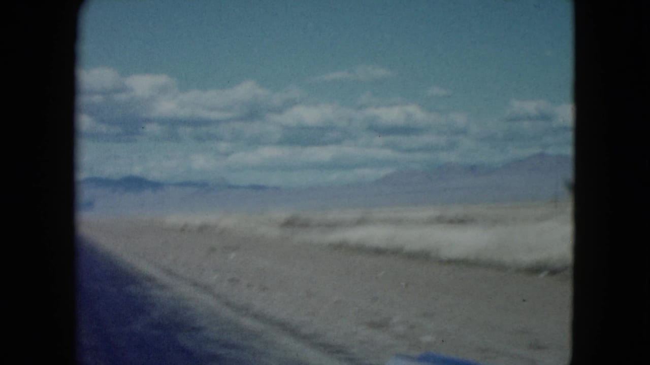 1959 8mm Nevada travel view with the edge of a desert highway, sandy terrain, distant hills, and scattered clouds.