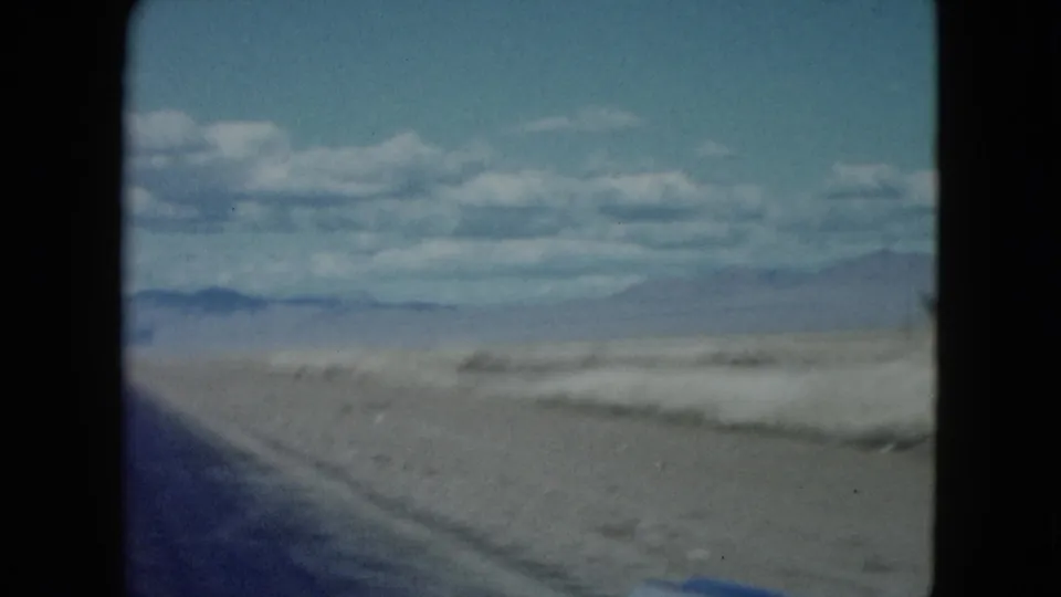 1959 8mm Nevada travel view with the edge of a desert highway, sandy terrain, distant hills, and scattered clouds.