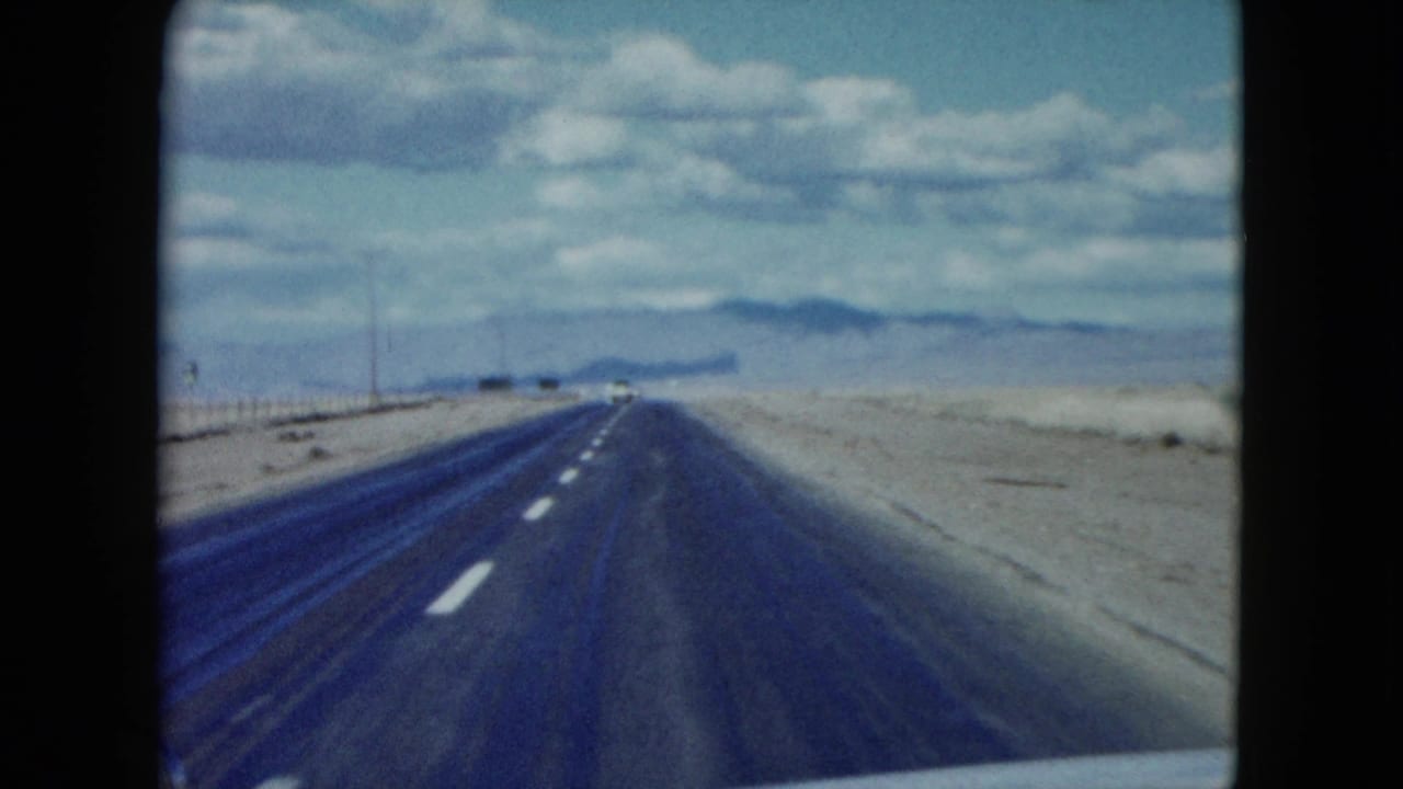 1959 8mm Nevada highway footage from a moving car, showing lane markings, open desert, roadside posts, and mountains ahead.
