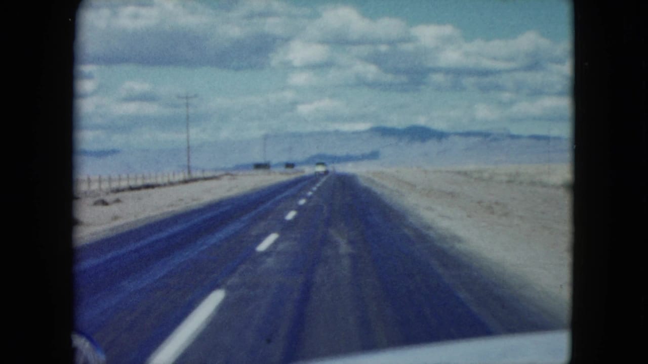 1959 8mm Nevada road view from a moving car, with a long highway, arid shoulders, distant mountains, and a small oncoming vehicle.