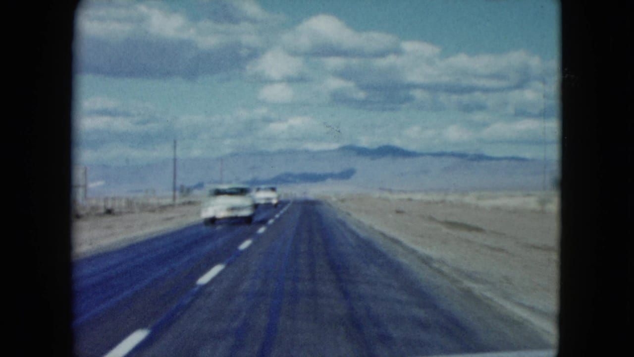 Two-lane desert highway receding toward distant mountains under a cloudy sky.