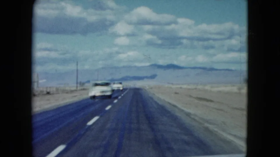 1959 Nevada road-trip footage from a moving car, looking down a two-lane desert highway toward low mountains and clouds.