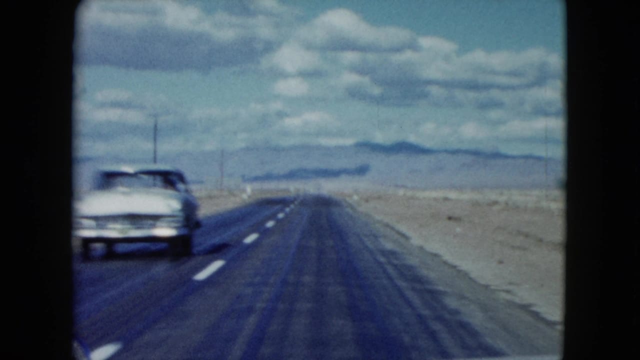 Light-colored car passes on a desert highway with mountains in the distance.