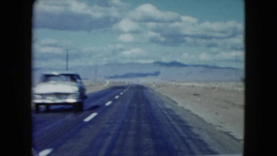 A light-colored period car passes on a Nevada desert highway in 1959, seen from a moving vehicle with open land beyond.