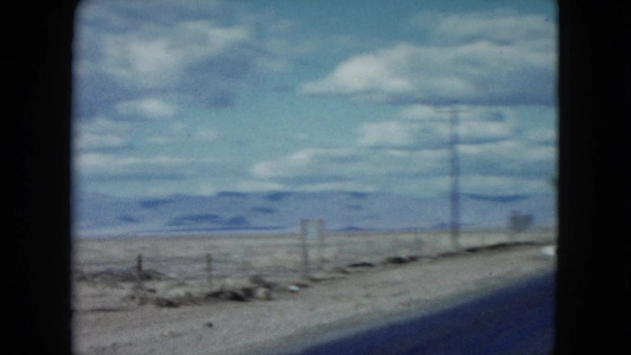 A side-looking view from the moving car shows Nevada desert roadside, utility poles, low mountains, and a small sign in 1959 home-movie footage.
