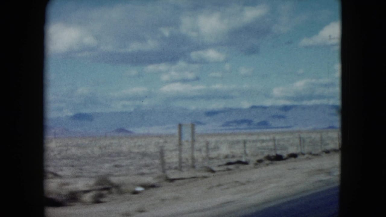 1959 Nevada desert roadside from a moving vehicle, with sparse scrub, fence posts, distant mountains, and a partly cloudy sky.