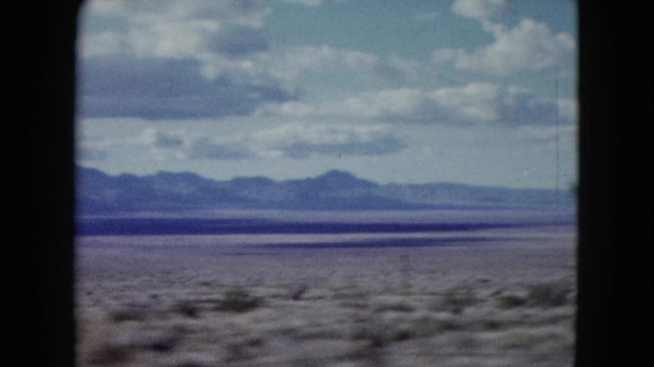Open desert plain and mountain range viewed through a dark car-window frame under clouds.