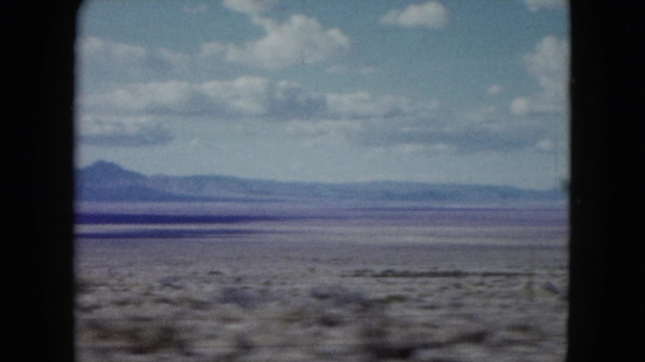 1959 Nevada desert landscape from a moving vehicle, with blurred scrub in front of a wide plain and mountain horizon.