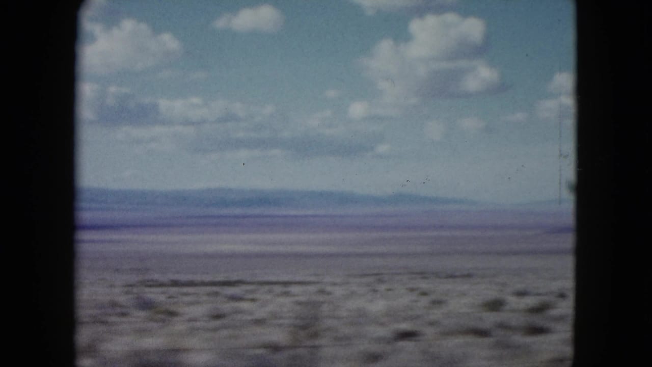 1959 Nevada desert plain seen from a moving car, with sparse scrub, a low horizon, distant hills, and partly cloudy sky.