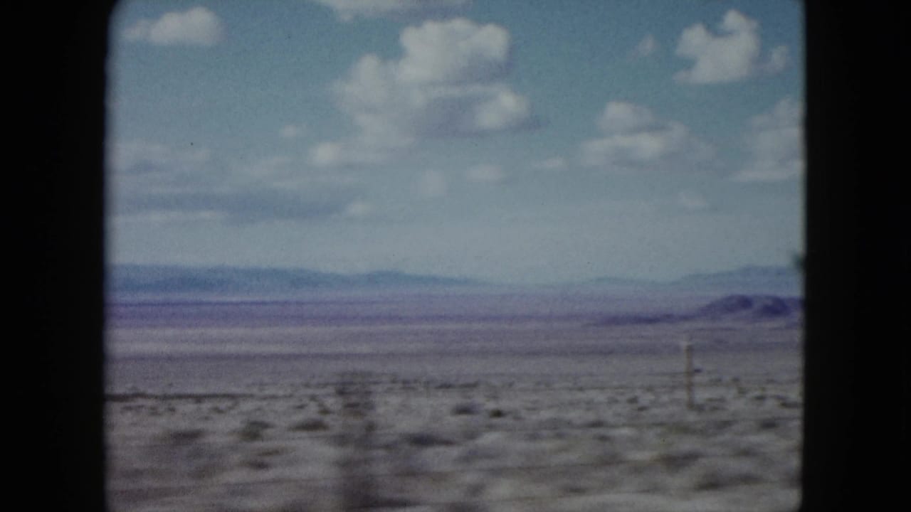 1959 Nevada 8mm frame showing an open desert plain, distant hazy mountains, scattered white clouds, and dark projector-like borders.