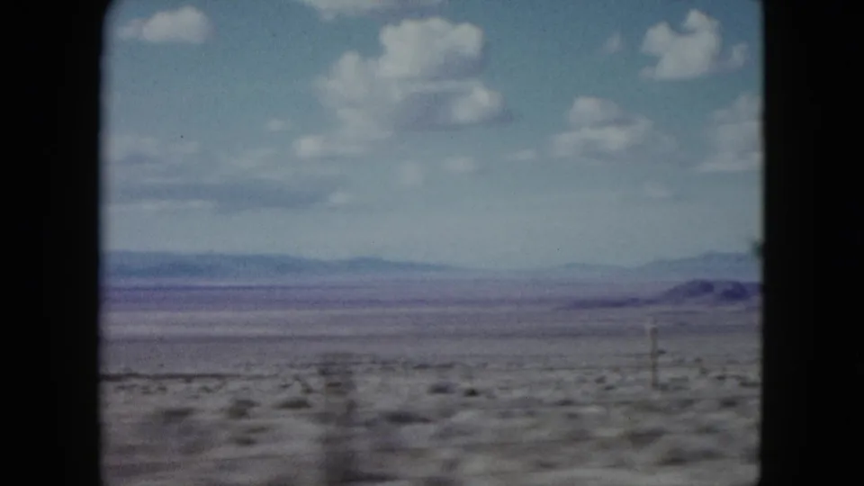 1959 Nevada 8mm frame showing an open desert plain, distant hazy mountains, scattered white clouds, and dark projector-like borders.