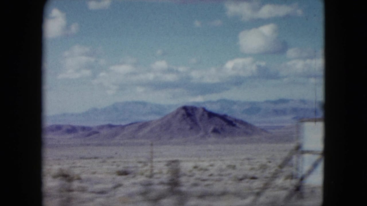 1959 Nevada 8mm frame showing a rounded desert hill, scrub-covered foreground, utility poles, distant ridges, and soft clouds.