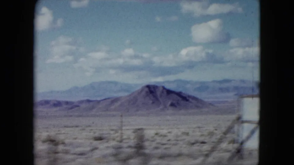 1959 Nevada 8mm frame showing a rounded desert hill, scrub-covered foreground, utility poles, distant ridges, and soft clouds.