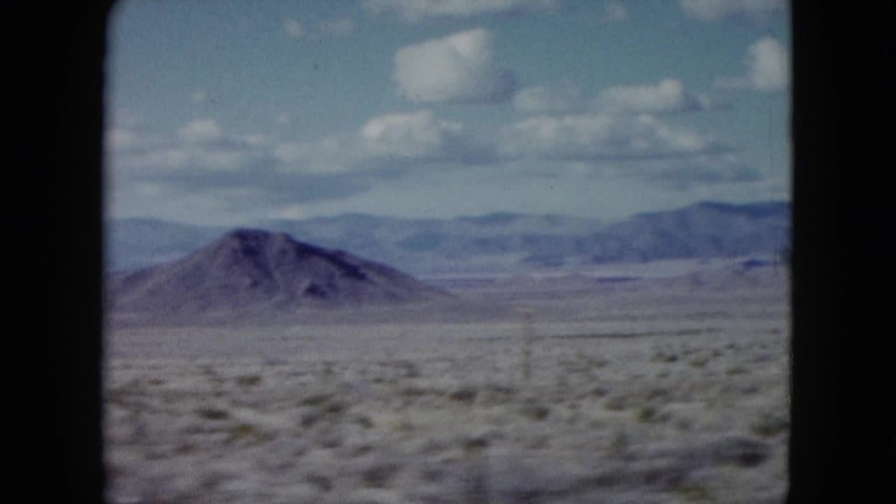 1959 Nevada desert landscape with a rounded hill, wide scrub plain, distant ridges, atmospheric haze, and visible 8mm frame edges.