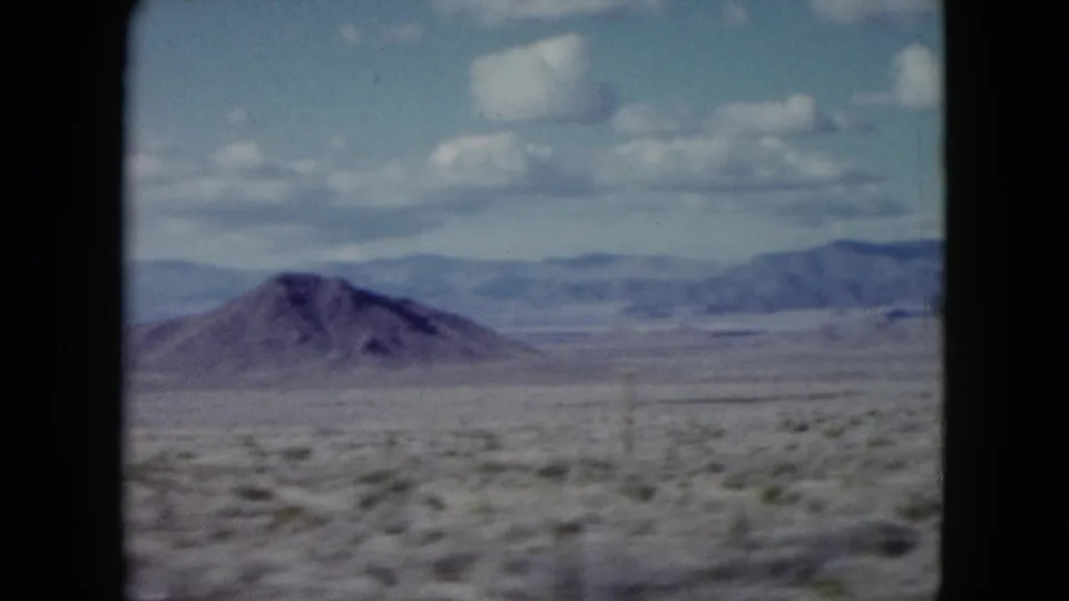 1959 Nevada desert landscape with a rounded hill, wide scrub plain, distant ridges, atmospheric haze, and visible 8mm frame edges.