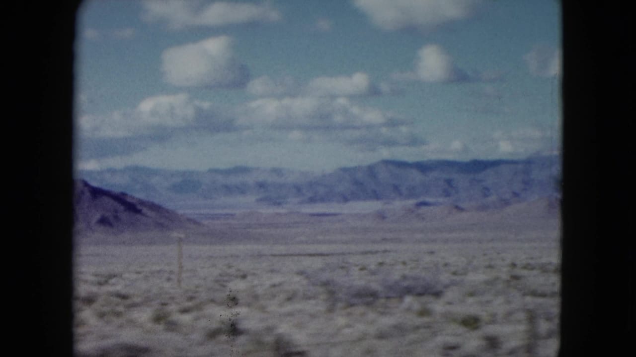 1959 Nevada 8mm travel frame showing dry desert scrub, distant mountains, and scattered white clouds seen through dark film borders.
