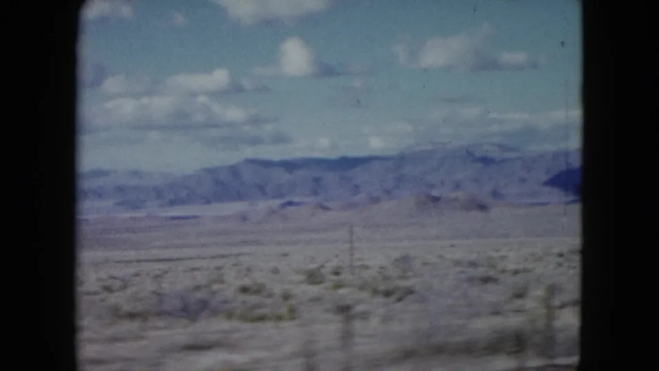 1959 Nevada home-movie frame of a broad arid plain with low vegetation, mountain ridges, and a partly cloudy blue sky.