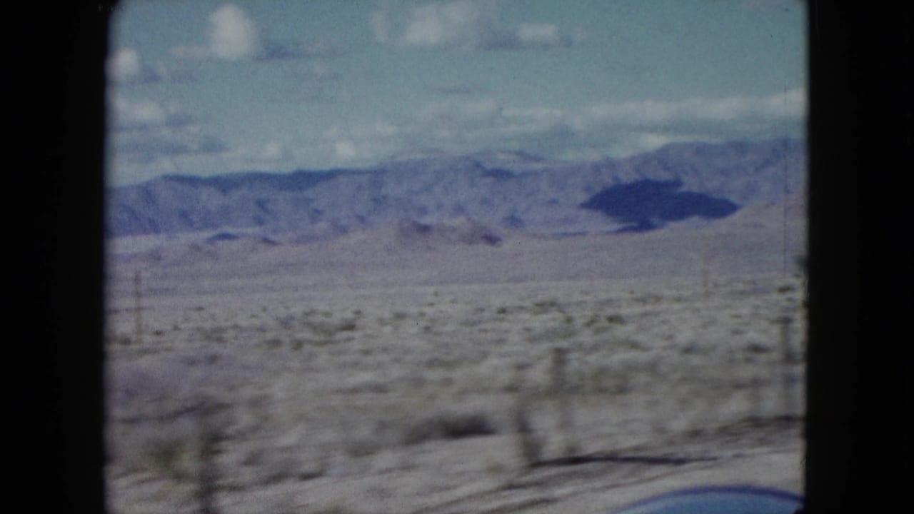 1959 Nevada 8mm frame with motion-blurred roadside scrub, fence posts, a dry valley, and distant mountains beneath clouds.