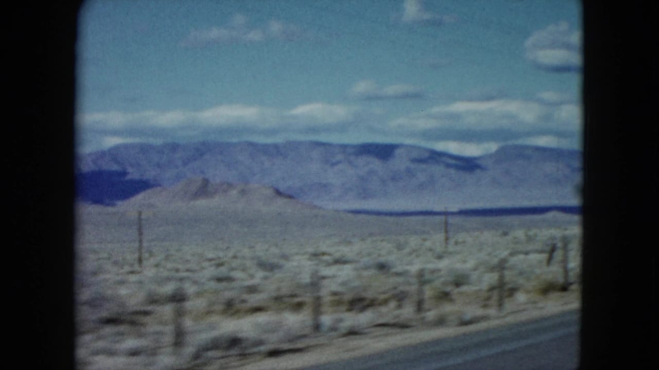 1959 Nevada travel frame showing a road edge beside desert scrub, fence posts, and a long mountain range under blue sky and white clouds.