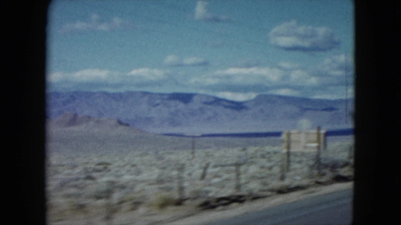 1959 Nevada 8mm frame showing an arid roadside field, fence posts, distant mountains, and a bright sky with scattered clouds.