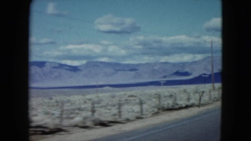 1959 Nevada travel view from a moving vehicle, with a roadside fence, sparse desert vegetation, mountains, and clouds.