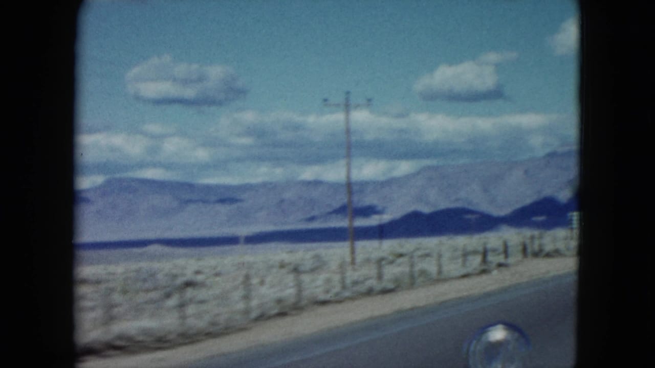 1959 Nevada 8mm roadside frame with utility poles, a fence line, open desert ground, and distant mountains under clouds.