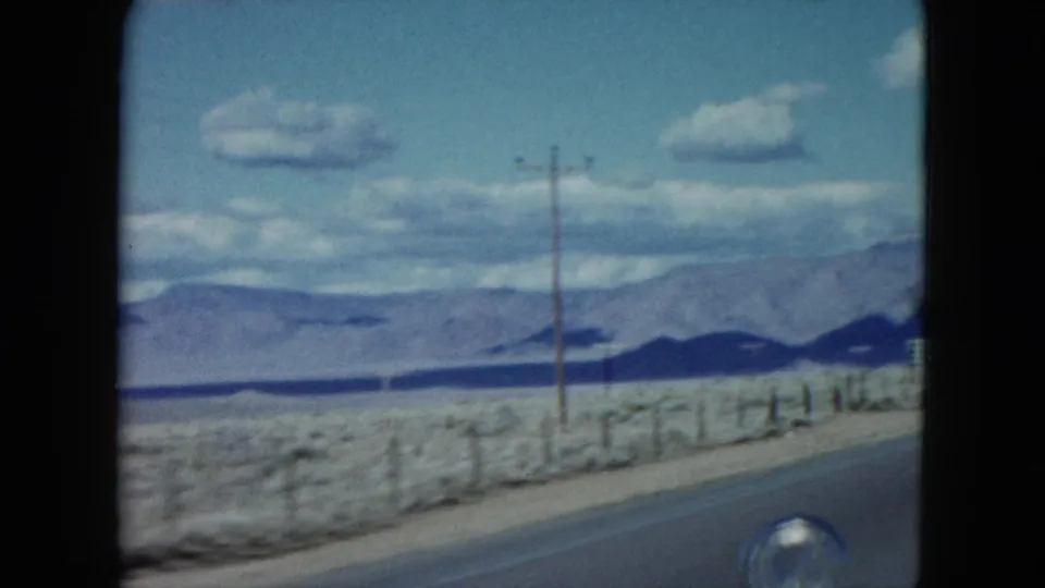 1959 Nevada 8mm roadside frame with utility poles, a fence line, open desert ground, and distant mountains under clouds.