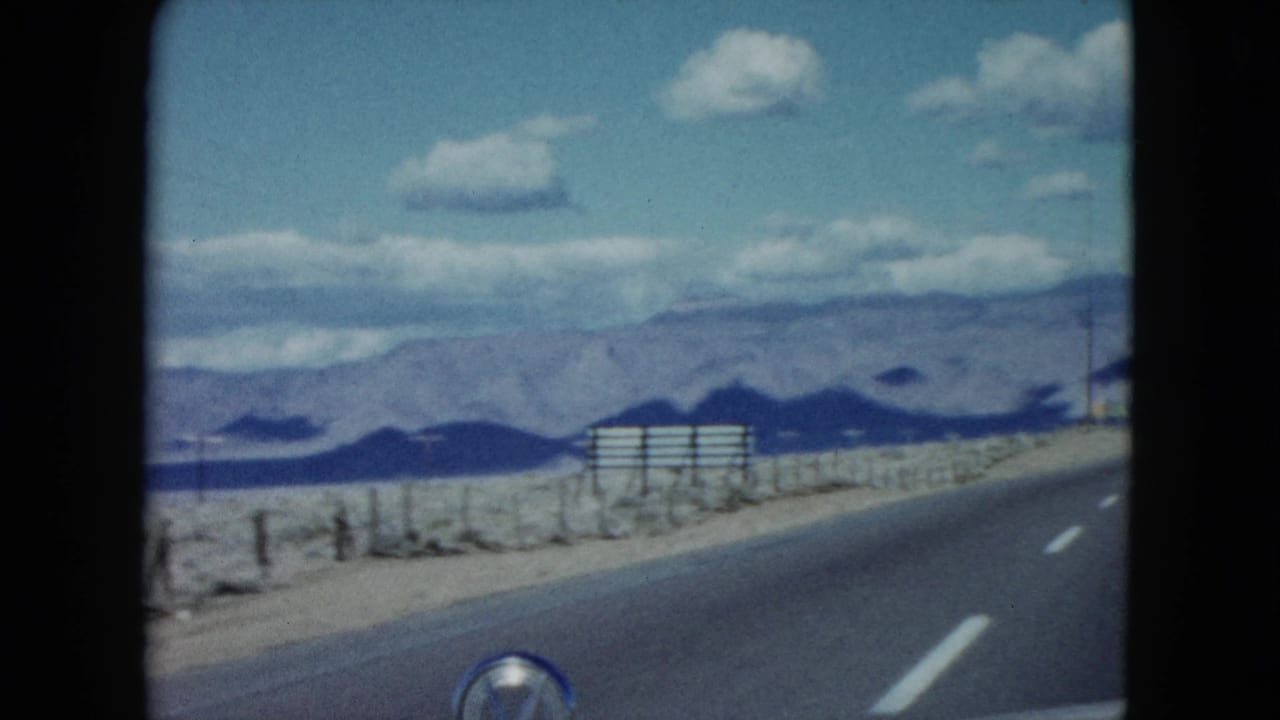 1959 Nevada road scene from a moving vehicle, showing lane markings, desert field, distant mountains, utility poles, and a small roadside sign.