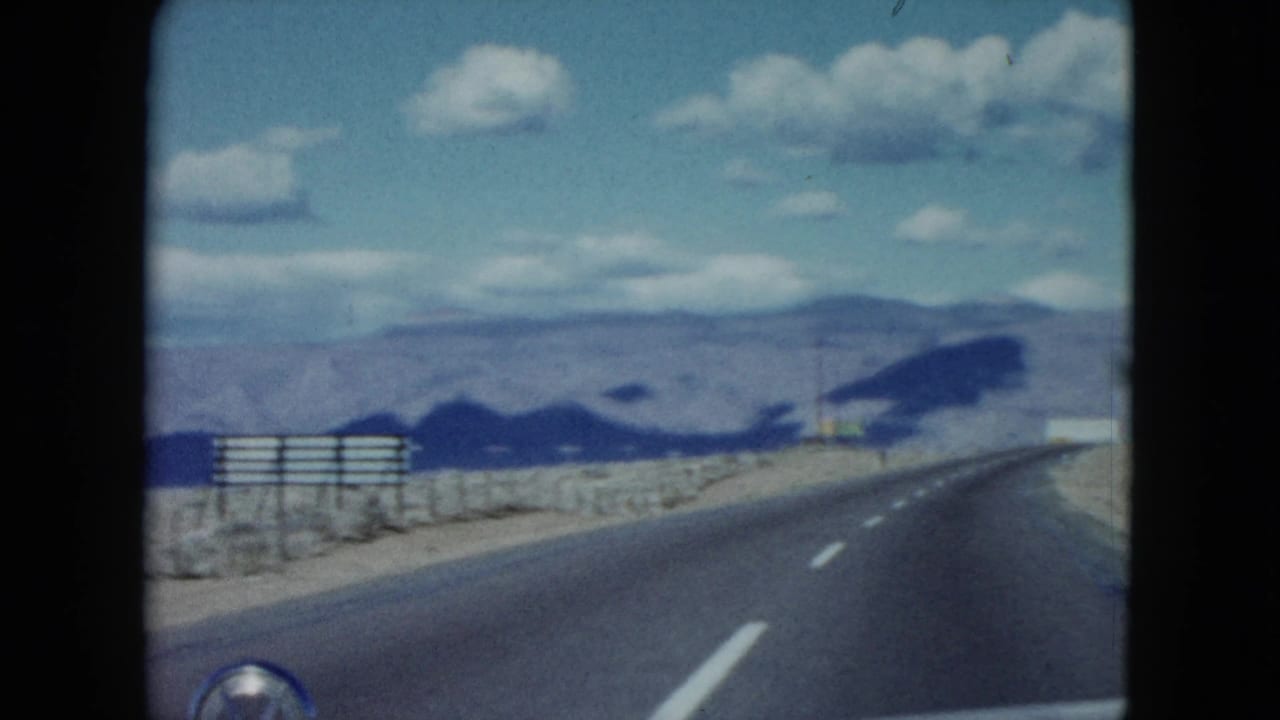 Desert highway seen from inside a moving car, with mountains and clouds ahead.