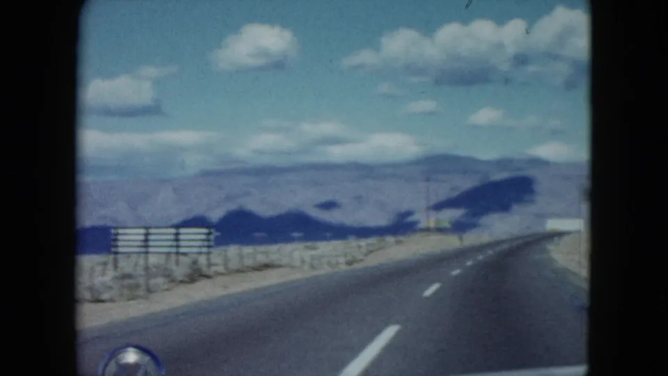 1959 Nevada 8mm road-trip view from a car, looking along a paved desert road toward low mountains under scattered clouds.