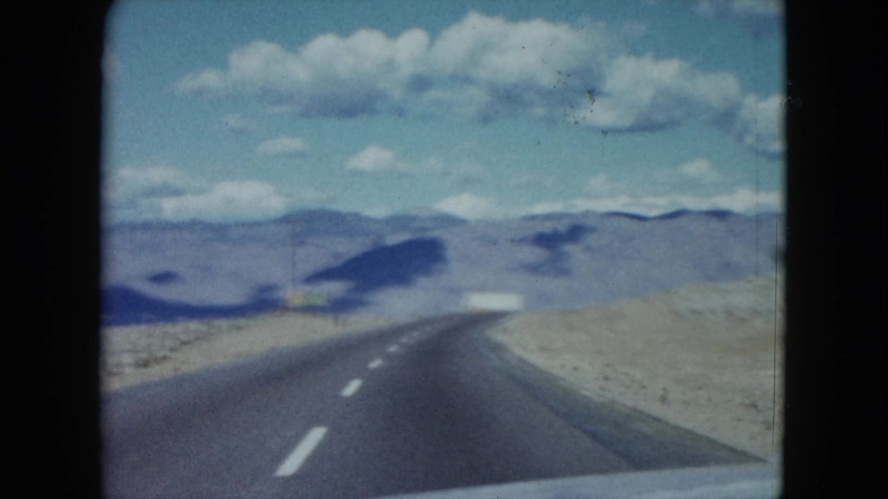 1959 Nevada home-movie frame of an open asphalt road through arid terrain, filmed from inside a moving vehicle.