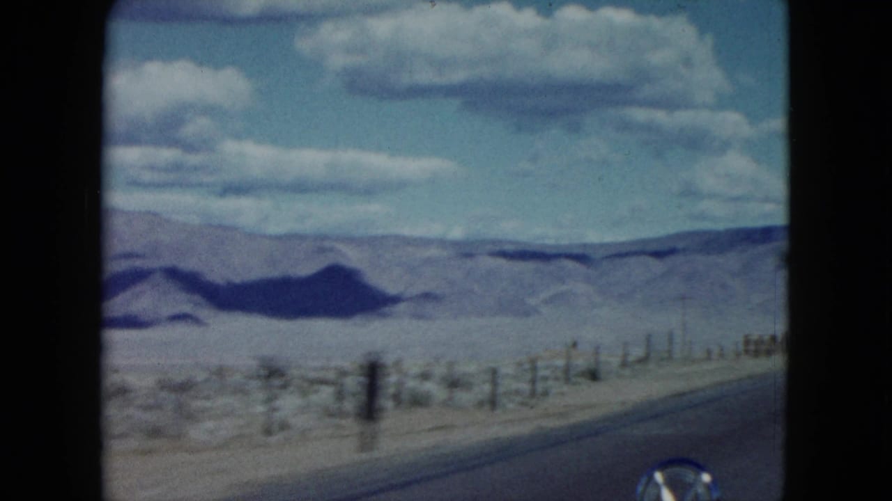 Nevada desert landscape from a moving 1959 road trip, showing roadside fencing, scrubland, and mountains beyond the highway.