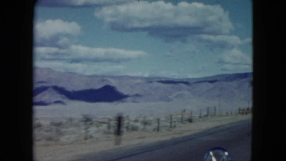 Nevada desert landscape from a moving 1959 road trip, showing roadside fencing, scrubland, and mountains beyond the highway.